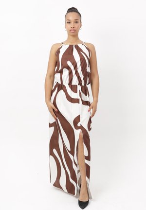 Mujer de pie, vestida con un vestido largo sin mangas con patrón marrón y blanco, con una abertura frontal y zapatos negros puntiagudos, sobre un fondo blanco liso.