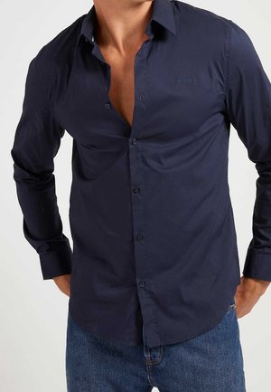 Hombre que lleva una camisa azul marino abotonada con los botones superiores desabrochados y jeans azules, con las manos apoyadas en las caderas.