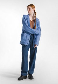 Lichtblauwe cardigan met ribgebreide manchetten en twee voorzakken, gelaagd over een bruine top, gecombineerd met losse blauwe denim broek en zwarte schoenen.