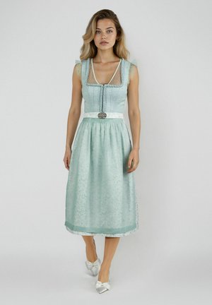 Junge Frau in einem hellblauen traditionellen Kleid mit Spitzenärmeln und weißen Ballerinas mit Schleife vor einem schlichten hellgrauen Hintergrund.