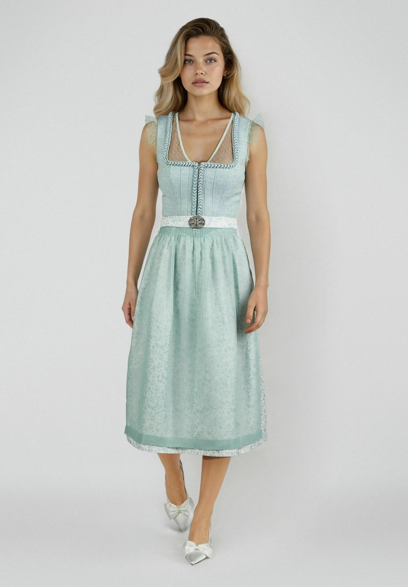 Junge Frau in einem hellblauen traditionellen Kleid mit Spitzenärmeln und weißen Ballerinas mit Schleife vor einem schlichten hellgrauen Hintergrund.