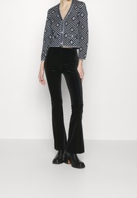 Pantalones acampanados de terciopelo negro combinados con un cárdigan corto y estampado que presenta diseños geométricos en negro, blanco y azul, terminado con botones.