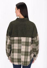 Camisa de pana verde oliva con una sección inferior de cuadros en crema y verde oscuro. Presenta un cuello tipo camisa y un corte holgado.