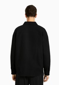 Svart långärmad skjorta med texturerad tyg, en traditionell krage och knappade manschetter. Har en avslappnad passform och rak skärning.