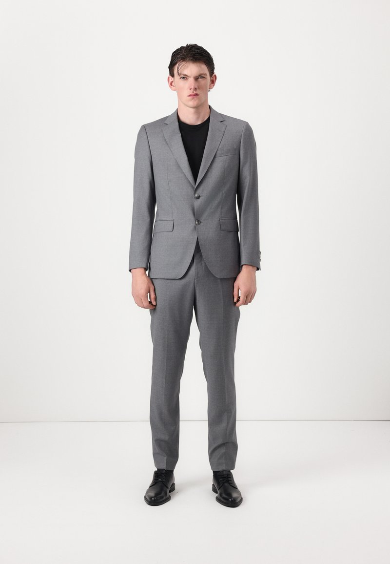 Grau Anzug mit zwei Knöpfen, mit Revers und seitlichen Taschen. Schwarzes Hemd darunter, kombiniert mit schwarzen LederSchuhen. Glatte Textur.