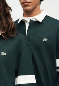 Deux personnes portant des maillots de rugby vert foncé avec des cols blancs et des bandes blanches sur les manches, arborant un petit logo de crocodile vert.