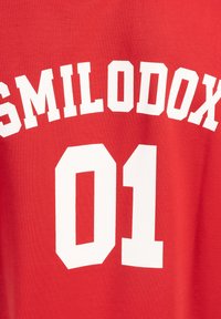 Camiseta de algodón roja con texto blanco en negrita: "SMILODON" encima de "01", con una fuente atlética clásica. Textura suave y diseño con cuello redondo.