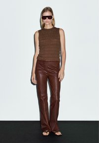 Top senza maniche marrone a costine con dettagli testurizzati, abbinato a pantaloni in pelle marrone aderenti. Modella con occhiali da sole oversize e sandali con punta aperta.