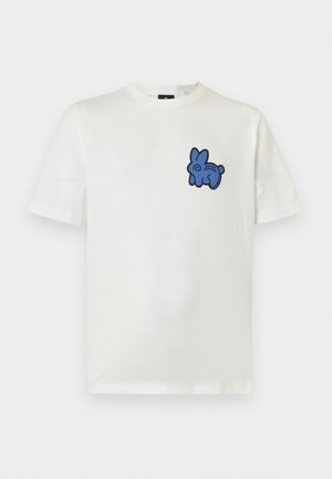 Ung mand med krøllet hår iført en hvid T-shirt med en blå kanin-illustration, grå plisserede bukser, hvide sneakers, stående med hænderne i lommerne.