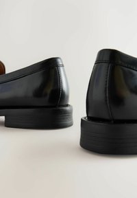 Zapatos de charol negro con un acabado suave, tacones bajos y detalles de costura sutiles. Punta redondeada y ligero elevamiento del tacón.