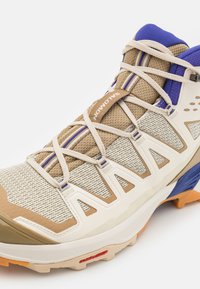 Beige terrengsko med mesh-overdel, teksturerte detaljer og blå høydepunkter. Har tykke snører, polstret krage og en robust såle.