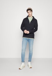 Zwarte rits sweatshirt over een lichtgroen t-shirt, gecombineerd met lichtblauwe jeans en grijze sneakers. Effen achtergrond, relaxte pasvorm.