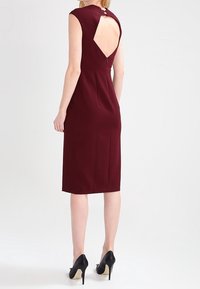 Bordeaux kleurige mouwloze jurk met een sleutelgatdetail aan de achterkant en een getailleerd ontwerp, tot aan de knie. Gemaakt met zwarte hakken.