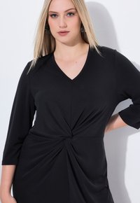 Vestido negro con escote en V, mangas tres cuartos y detalle de nudo en la cintura, fabricado con una tela suave y textura drapeada.
