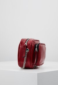 Borsa a tracolla rossa in pelle sintetica con motivo coccodrillo testurizzato, doppie zip e tracolla in catena argentata. Forma compatta e rettangolare.