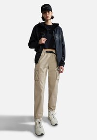 Zwarte windjack met capuchon, cropped zwart T-shirt, beige cargo broek met zakken, en witte sneakers met zwarte zolen. Zwarte pet.