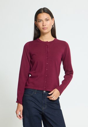 Femme aux cheveux lisses et foncés porte un cardigan bordeaux boutonné et un jean bleu foncé, debout devant un fond clair uni.