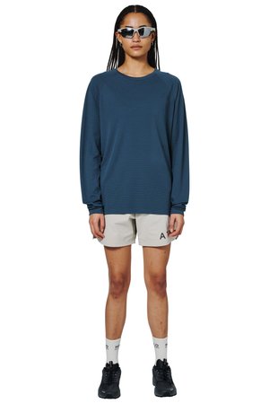 Person iført mørkeblå langærmet skjorte, beige shorts med tekst, hvide sokker, sorte sneakers og mørke solbriller, der står oprejst.