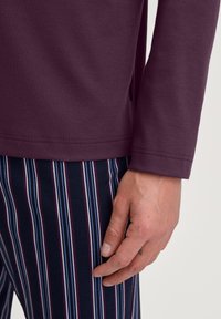 Camicia a maniche lunghe in cotone viola scuro. Mano appoggiata su pantaloni a righe blu navy con linee verticali rosse, blu e bianche.