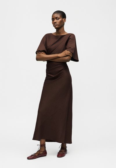 Γυναίκα με κοντά μαλλιά στέκεται με σταυρωμένα χέρια, φορώντας καφέ φόρεμα και κόκκινα δερμάτινα παπούτσια με κορδόνια, μπροστά σε απλό φωτεινό φόντο.