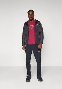 Juoda pūkinė striukė ant bordo spalvos marškinėlių su užrašu „Peak Performance“, derinama su juodomis kelnėmis ir juodais žygių batais.