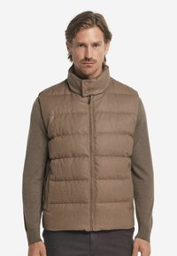 Braune gestepptes Weste mit hohem Kragen und Reißverschluss. Verfügt über eine glatte Textur und Druckknöpfe am Kragen. Getragen über einem braunen Langarmshirt.