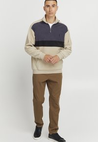 Suéter beige de media cremallera con rayas azul marino y negras, combinado con pantalones marrones y zapatillas negras, que cuenta con un bolsillo frontal.