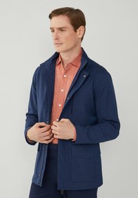 Chaqueta azul oscuro hecha de una tela texturizada con un cuello alto, bolsillos frontales y cierre de cremallera, llevada sobre una camisa de botones color naranja claro.