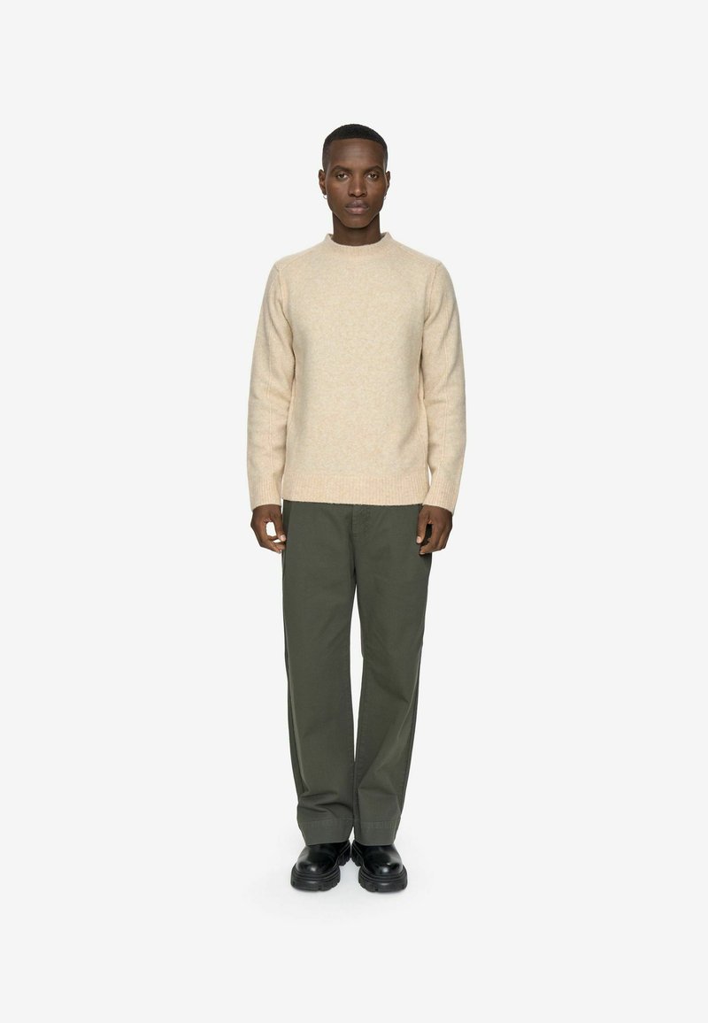 Un pull en tricot beige avec un col rond, associé à un pantalon ample vert olive et des chaussures noires. L'ensemble présente une apparence texturée.