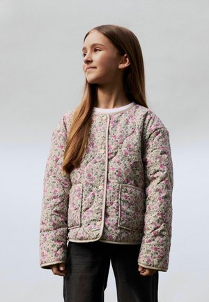 Jeune fille aux longs cheveux portant une veste matelassée légère à motif floral avec des poches devant, regardant avec confiance sur le côté sur un fond uni.
