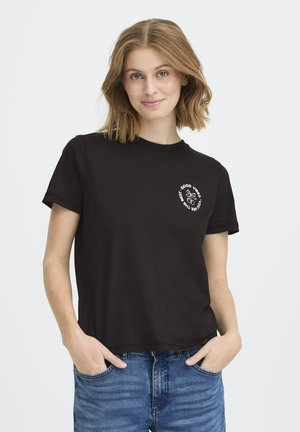 Junge Frau mit schulterlangem, hellbraunem Haar trägt ein schwarzes T-Shirt mit dem Aufdruck "Good vibes, you are the best" und einem Schmetterlingsgrafik sowie blaue Jeans.