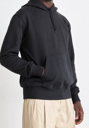 Man som bär en mörkgrå hoodie med en framficka och beiga veckade byxor, med handen vila i fickan, mot en neutral bakgrund.