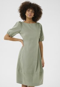 Kratka obleka z napihnjenimi rokavi v mahasto zeleni bombažni tkanini, ki ima ravno silhueto, kvadratni izrez in stranske žepke. Gladek, lahkoten tekstur.