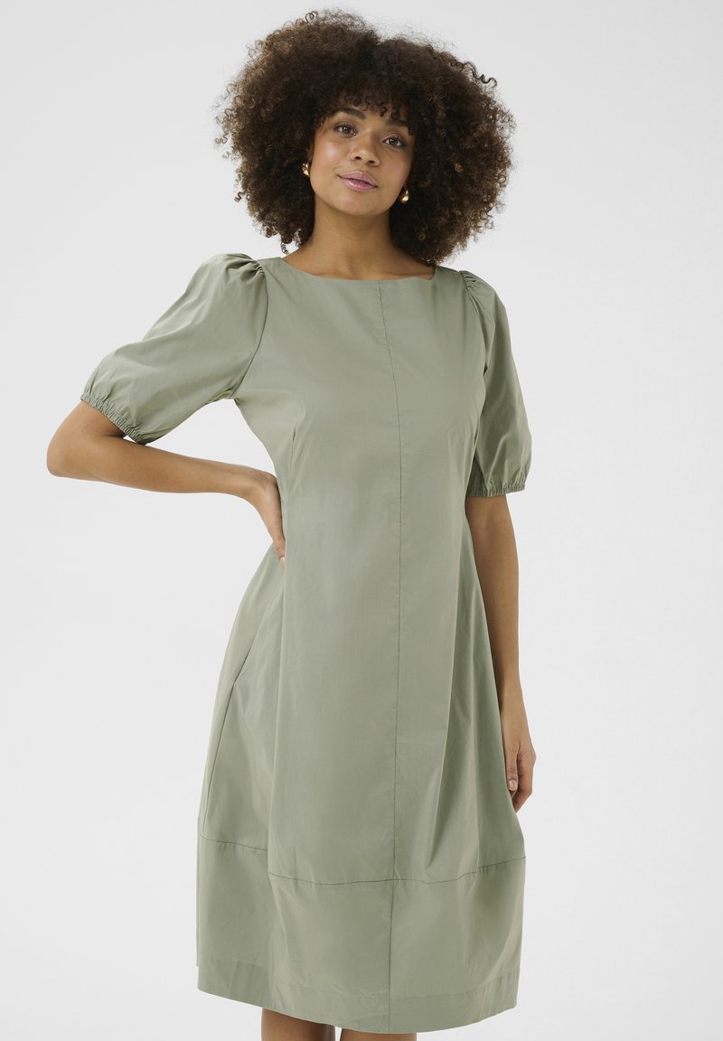 Kratka obleka z napihnjenimi rokavi v mahasto zeleni bombažni tkanini, ki ima ravno silhueto, kvadratni izrez in stranske žepke. Gladek, lahkoten tekstur.