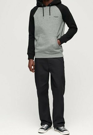 Homme portant un sweat à capuche noir et gris avec le logo « SuperDry », un pantalon noir et des baskets noir et blanc, debout avec une main dans la poche.