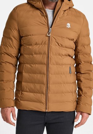 Homme portant une veste matelassée brune zippée avec capuche et un petit logo blanc sur la poitrine, assortie à un pantalon foncé.