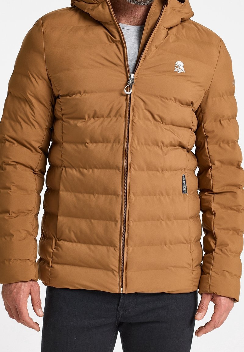 Homme portant une veste matelassée brune zippée avec capuche et un petit logo blanc sur la poitrine, assortie à un pantalon foncé.