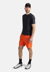 Svart kortärmad träningsskjorta, ljusorange shorts och grå sneakers med svarta accenter. Bär en svart keps med logotyp.