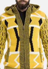 Gelbe Strickjacke mit Reißverschluss und schwarz-weißen geometrischen Mustern. Verfügt über einen strukturierten, gerippten Kragen und Zopfmuster an den Ärmeln.
