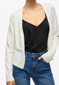 Cardigan bianco a maglia con apertura frontale, polsini e orlo ribattuti, indossato sopra un canottiera nera, abbinato a jeans blu a vita alta.