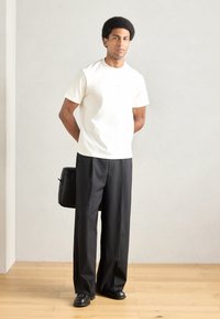 Biała koszulka z krótkim rękawem o luźnym kroju, zestawiona z czarnymi szerokimi spodniami. Czarna torba przewieszona przez ramię, stojąca na drewnianej podłodze.