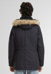 Črna parka jakna z umetnim krznom na kapuci. Ima teksturiran ovratnik, šive na prsih in stranske žepe. Ohlapen kroj s srednjo dolžino.