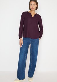 Langärmliges, dunkelviolettes Oberteil mit V-Ausschnitt und dekorativen Details, kombiniert mit weiten blauen Jeans und hellfarbenen Sneakers.