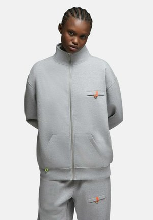 Personne aux cheveux tressés portant une veste gris ample zippée et un pantalon assorti avec de petits détails orange et marron sur les poches.