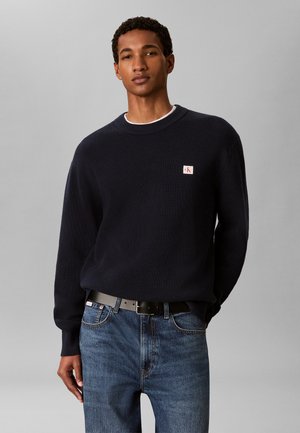 Giovane uomo che indossa un maglione nero lavorato a maglia con una piccola toppa logo sul petto e jeans blu con cintura nera.