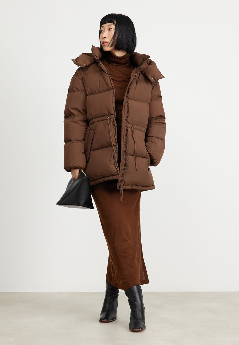 Brązowa ocieplana kurtka z wiązaniem w talii i kapturem, noszona na brązową sukienkę. Czarne botki na obcasie i mała czarna torebka dopełniają strój.