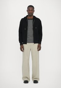 Felpa nera con zip sopra una camicia a righe orizzontali, abbinata a pantaloni larghi beige chiaro e scarpe nere, in piedi davanti a uno sfondo bianco.