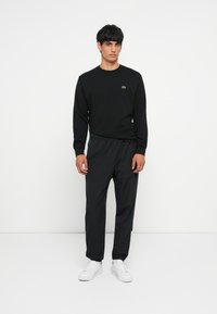 Black long-sleeve shirt with green logo, paired with dark pants with elastic cuffs, finished with white sneakers. Plain background.