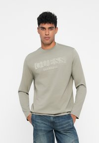 Tricou cu mânecă lungă, de culoare verde măslin deschis, cu un logo texturat „GUESS” pe piept, realizat din bumbac, cu un guler tip crew și o croială slim.