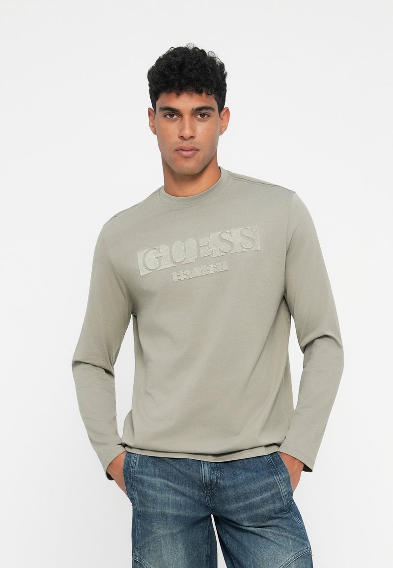 Tricou cu mânecă lungă, de culoare verde măslin deschis, cu un logo texturat „GUESS” pe piept, realizat din bumbac, cu un guler tip crew și o croială slim.